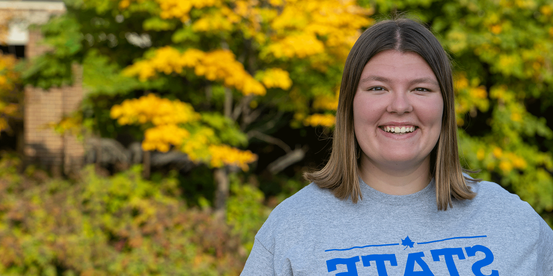 A white female student with shoulder-length brown hair poses outside, smiling. She wears a grey T-shirt with STATE in blue lettering on the front. Green trees with a variety of light yellow/orange leaves are blurred in the background.